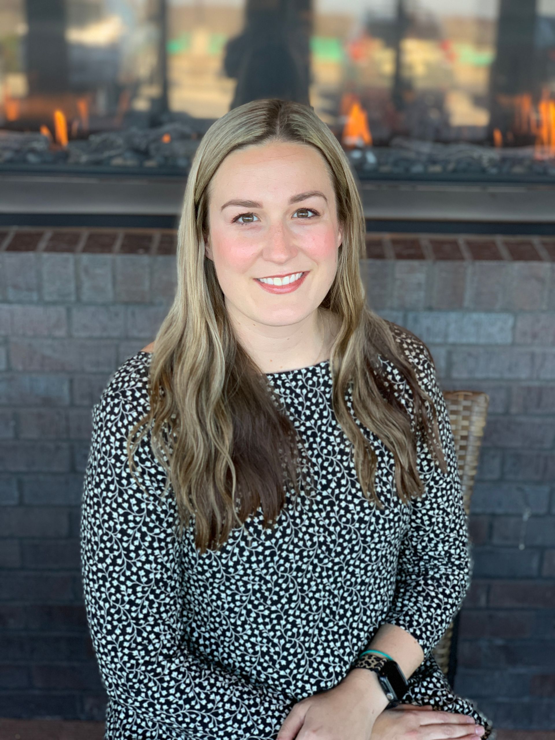 A smiling person with long light brown hair wearing a black and white patterned top, sitting in front of a brick fireplace.