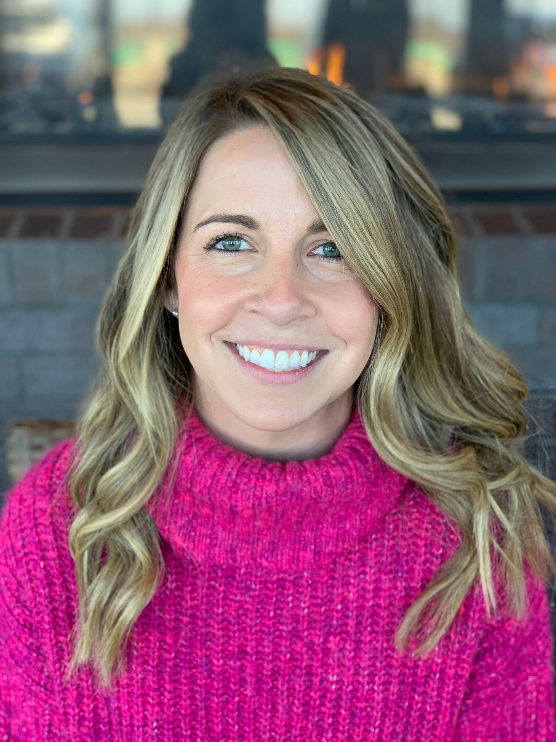 A woman with blonde hair and a bright pink turtleneck sweater smiling at the camera in a room with a brick fireplace.