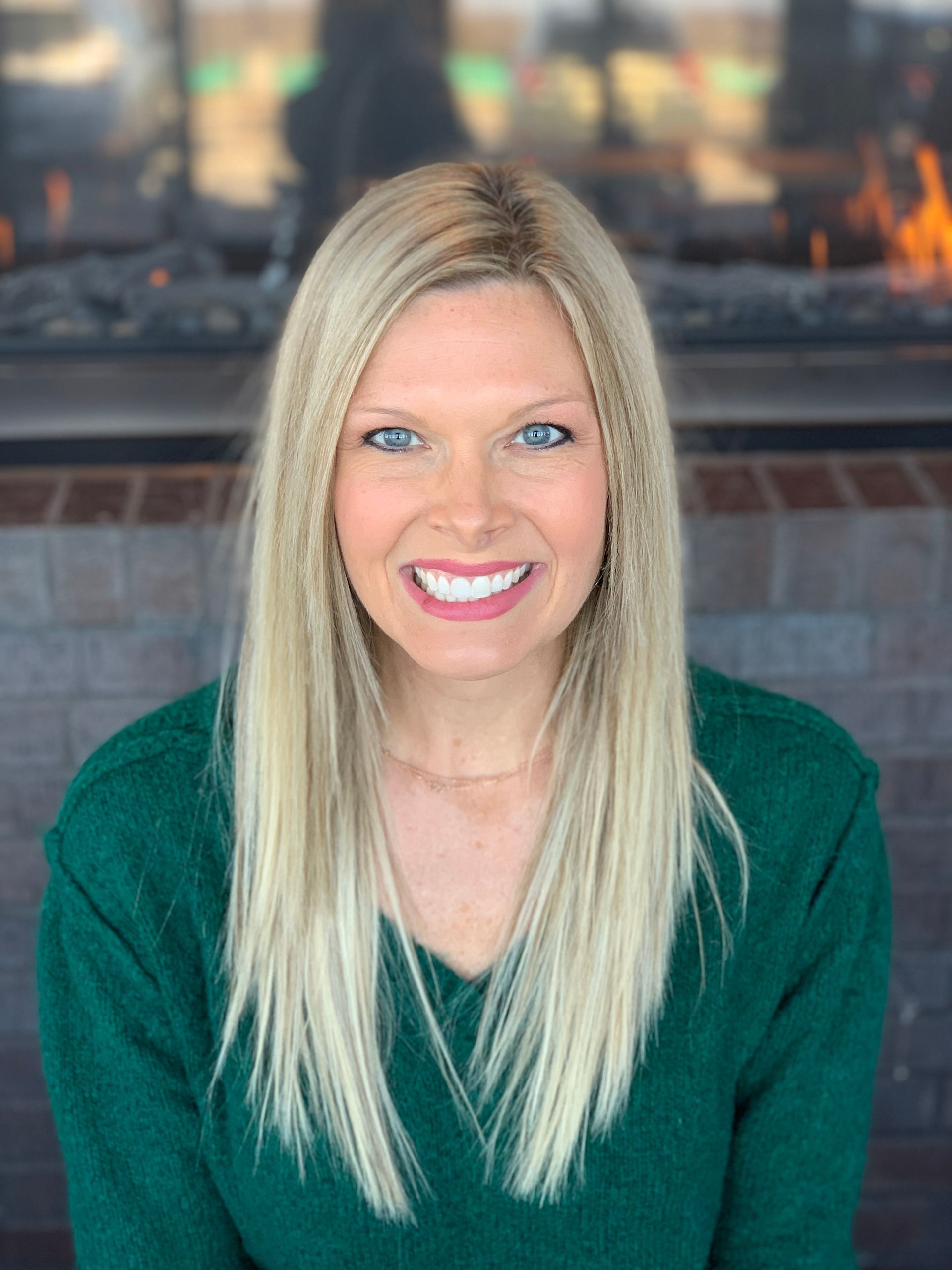 A smiling person with long blonde hair wearing a green sweater, standing in front of a brick fireplace.