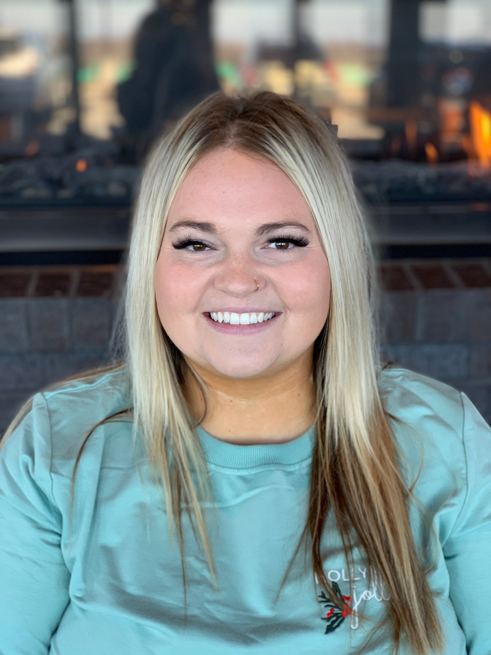 A smiling person with long blonde hair, wearing a light blue long-sleeved shirt, poses in front of a stone fireplace.