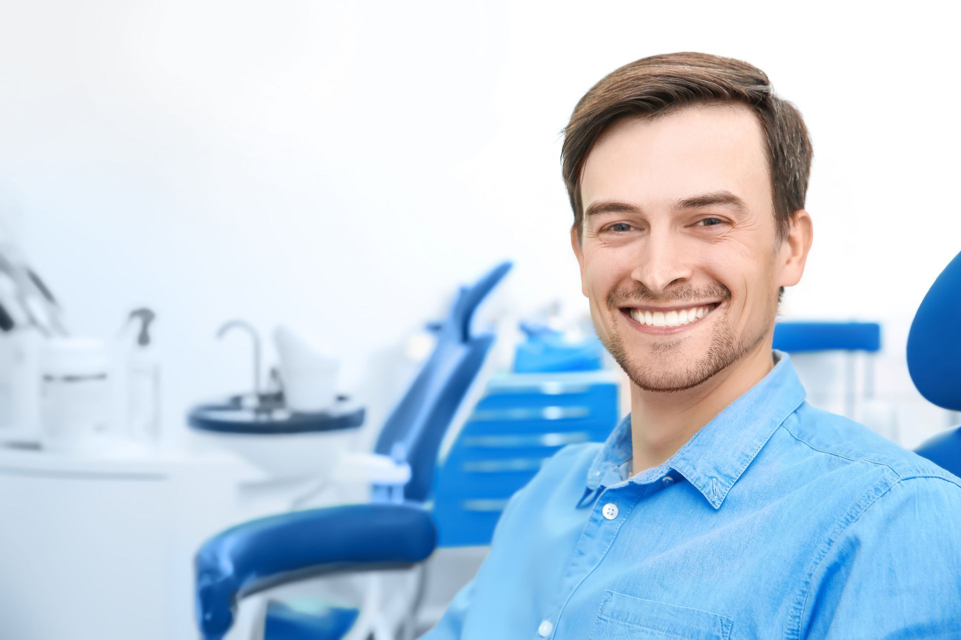 A smiling person in a blue shirt sits in a dental chair within a professional clinic setting.