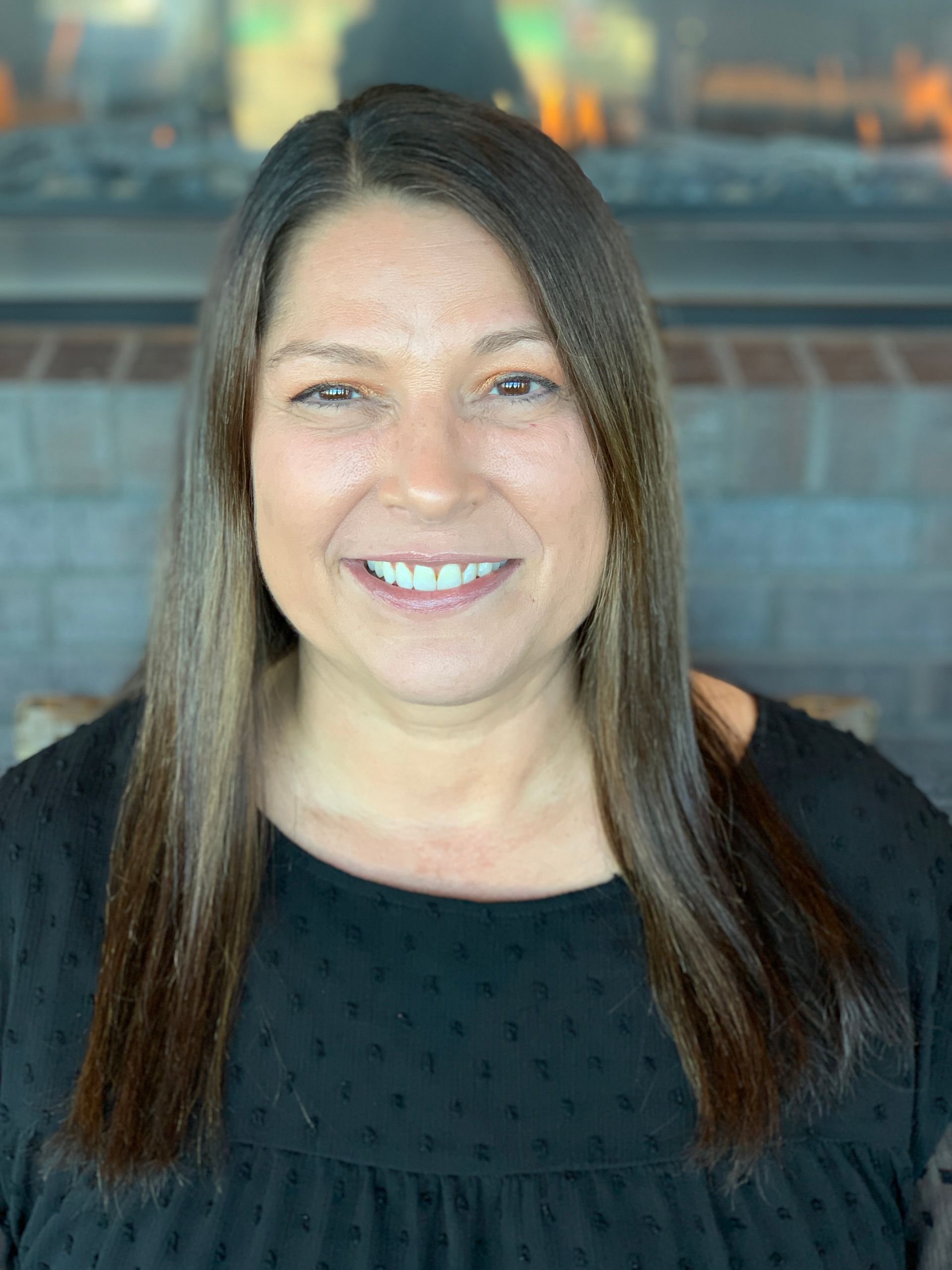 A person with long, dark hair and a warm smile poses in front of a brick fireplace.