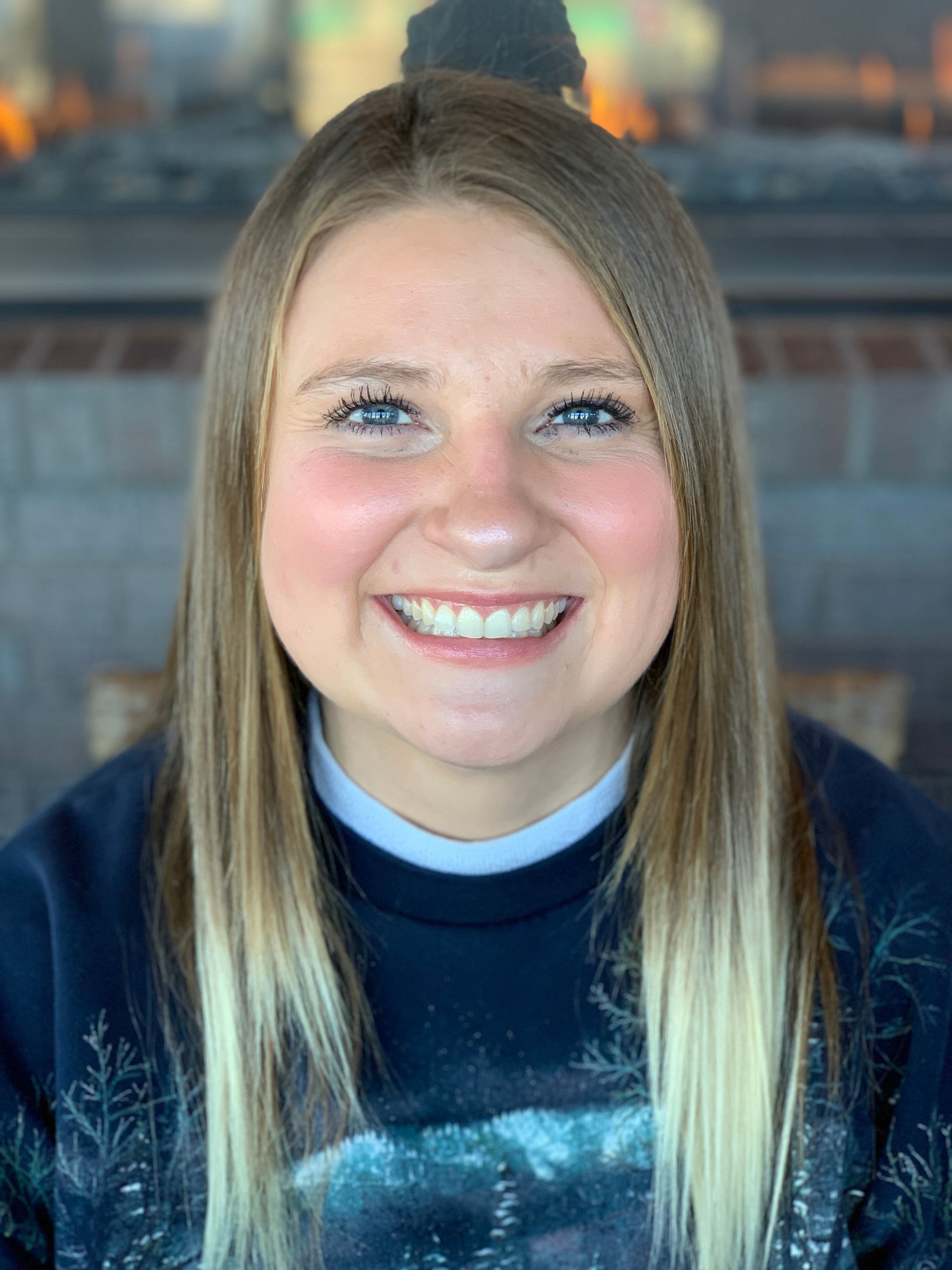 A smiling person with long, two-toned hair wearing a dark, patterned sweatshirt in front of a brick fireplace.