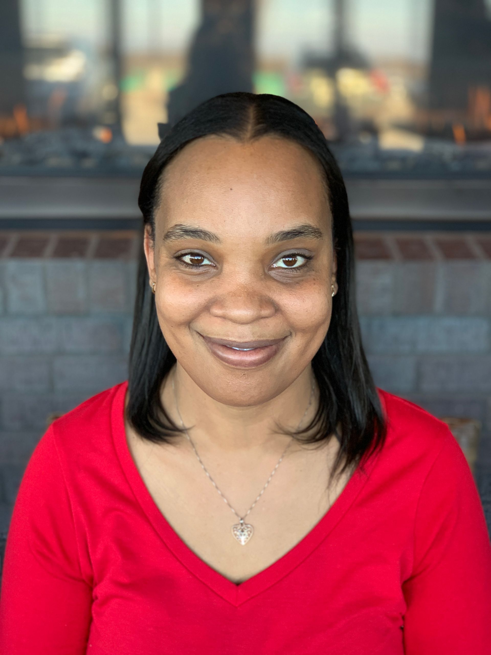 A person with dark hair wearing a red v-neck shirt and a silver necklace, smiling in front of a stone fireplace.