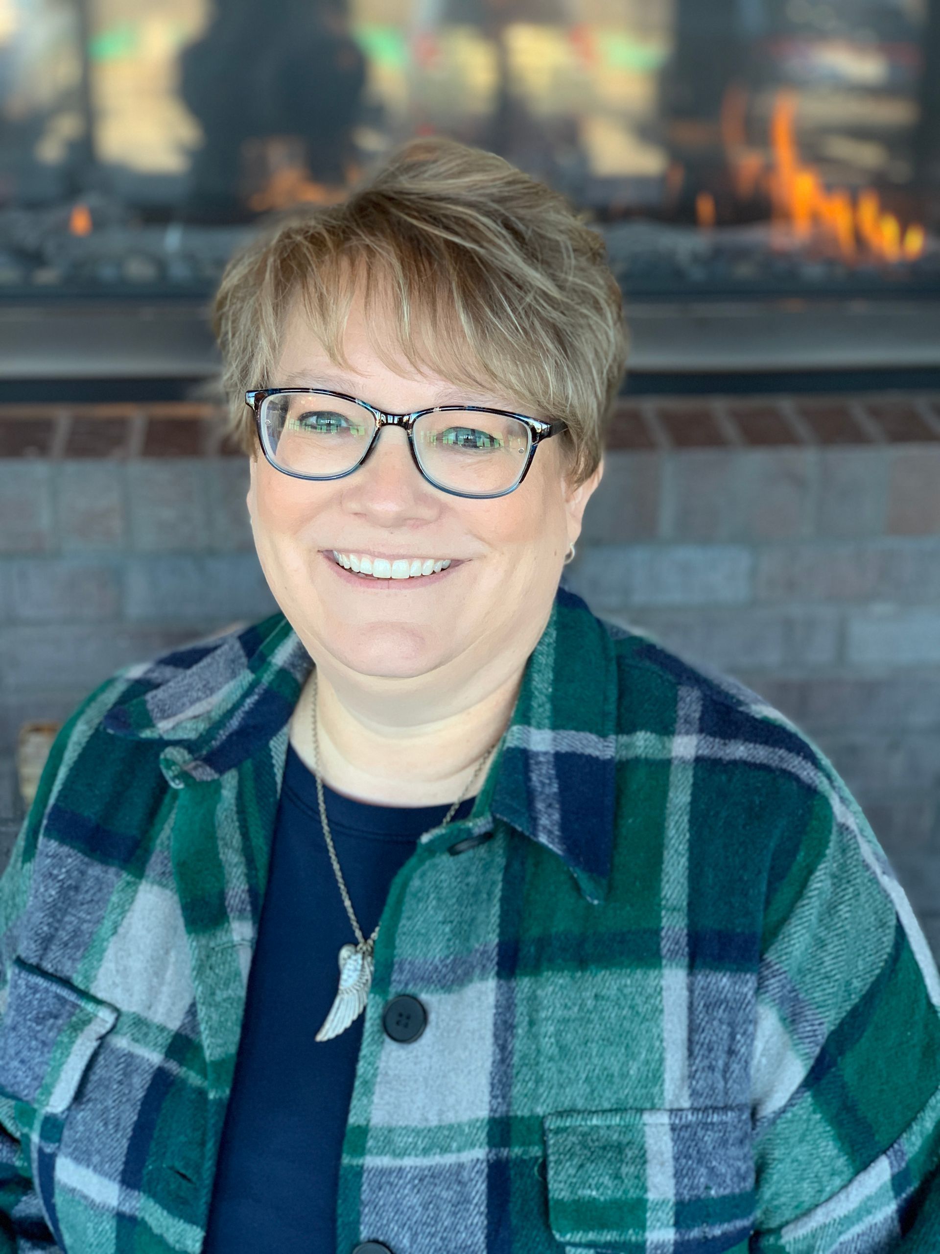 A person with short hair and glasses smiles while wearing a dark green, blue, and white plaid shirt in front of a fireplace.