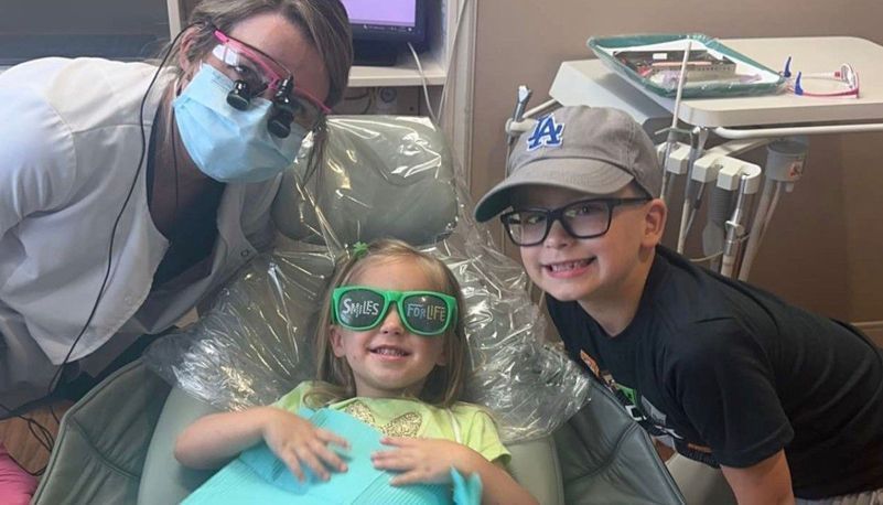 A dentist wearing a mask and loupes smiles beside a child in a dental chair wearing green glasses and a boy in a cap.