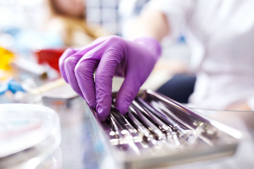 A person wearing purple latex gloves selects a metal dental instrument from a stainless steel tray in a medical clinic.