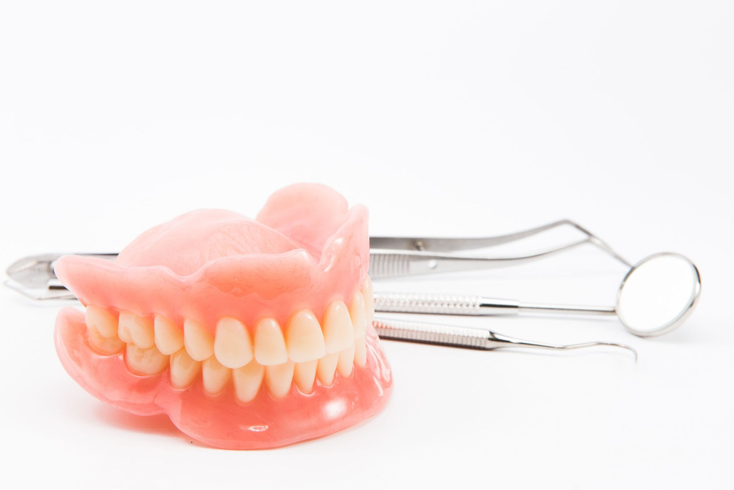 A full set of pink dentures resting on a white surface next to a dental mirror and a metal dental explorer tool.