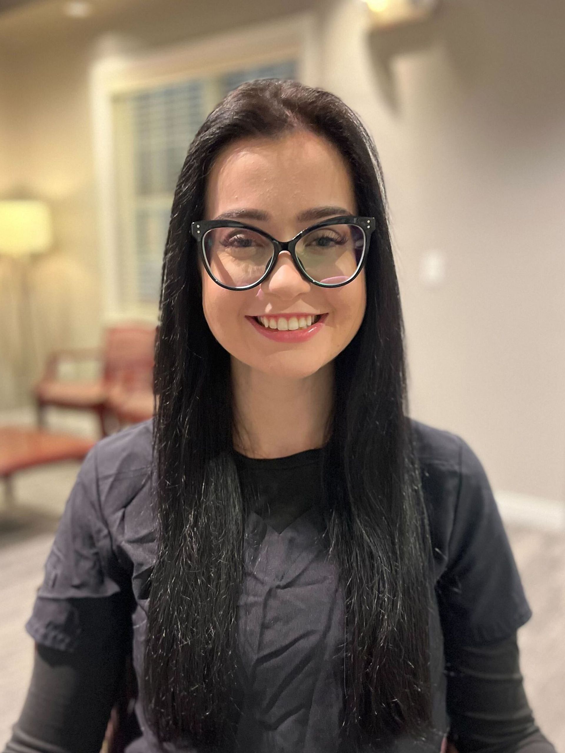 A smiling person with long dark hair and dark-rimmed glasses wearing a grey shirt, indoors with blurred room furniture.