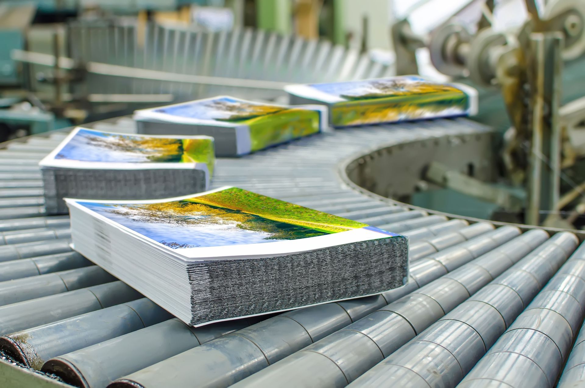 Stacks of printed books with colorful landscape covers on a factory conveyor belt.