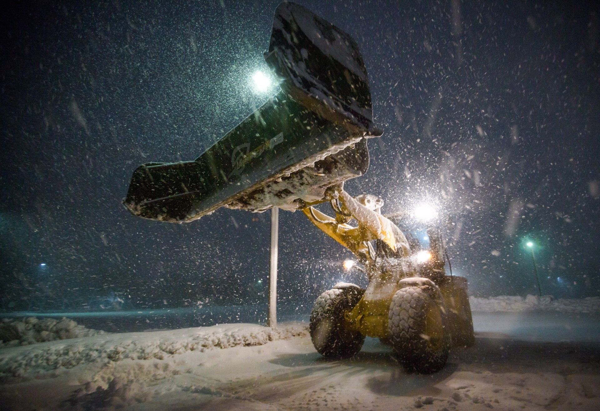 Yellow snowplow lifting snow in a nighttime snowstorm, with lights illuminating the scene.
