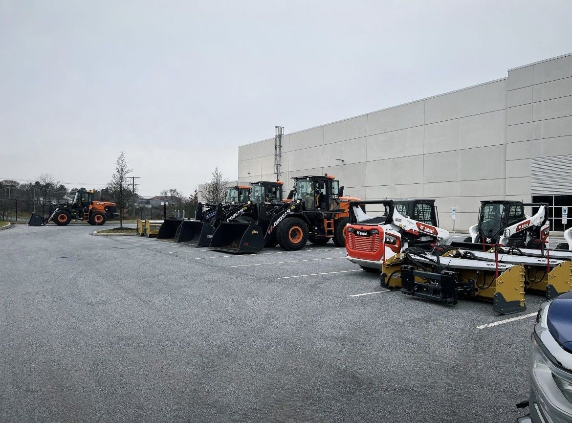 Several pieces of heavy machinery, like loaders and Bobcats, parked outside a large white building on a cloudy day.