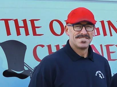 A man wearing a red hat and glasses stands in front of a sign that says the origin