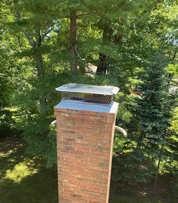 A brick chimney with a metal cover on top of it is surrounded by trees.