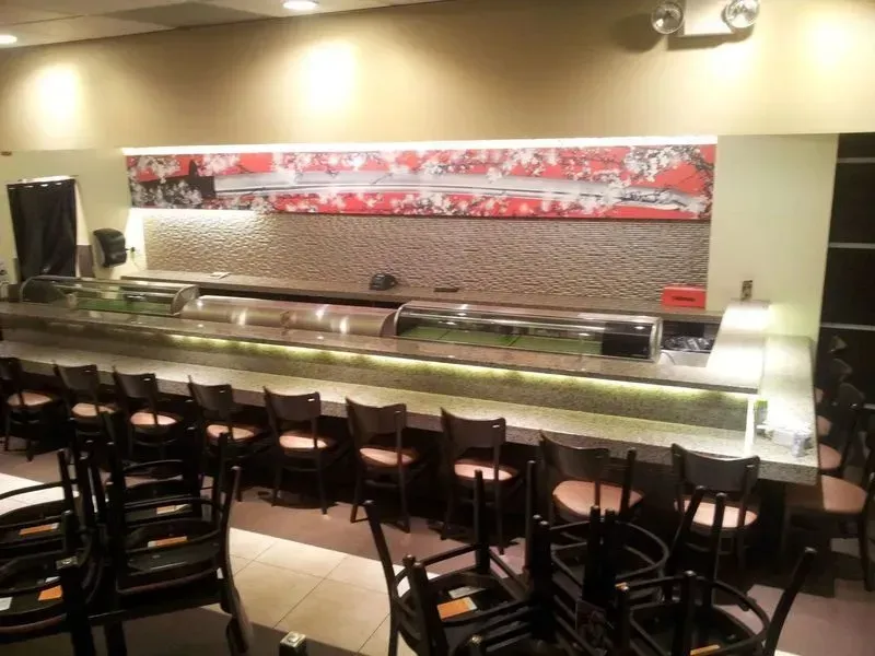 Sushi bar with seating; counter with display cases, red artwork, brown chairs.