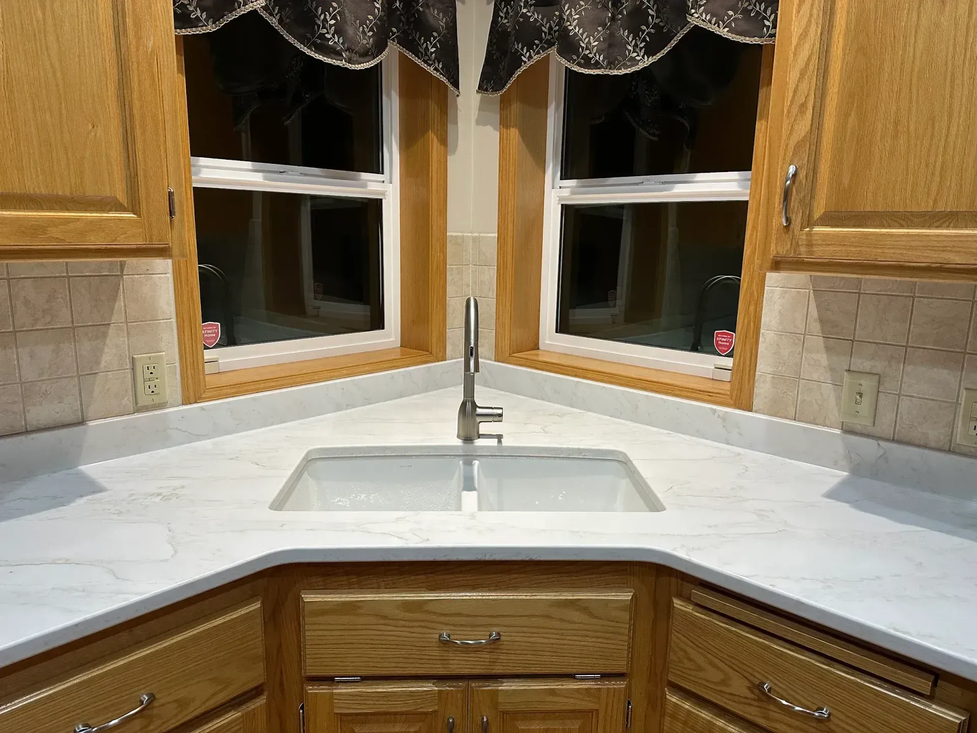 Kitchen corner with a white countertop, sink, and two windows above. Wooden cabinets are below.