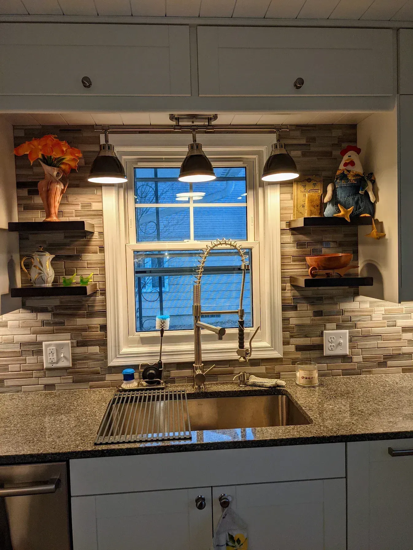 Kitchen sink with faucet, window, granite countertop, backsplash, floating shelves, and three pendant lights.