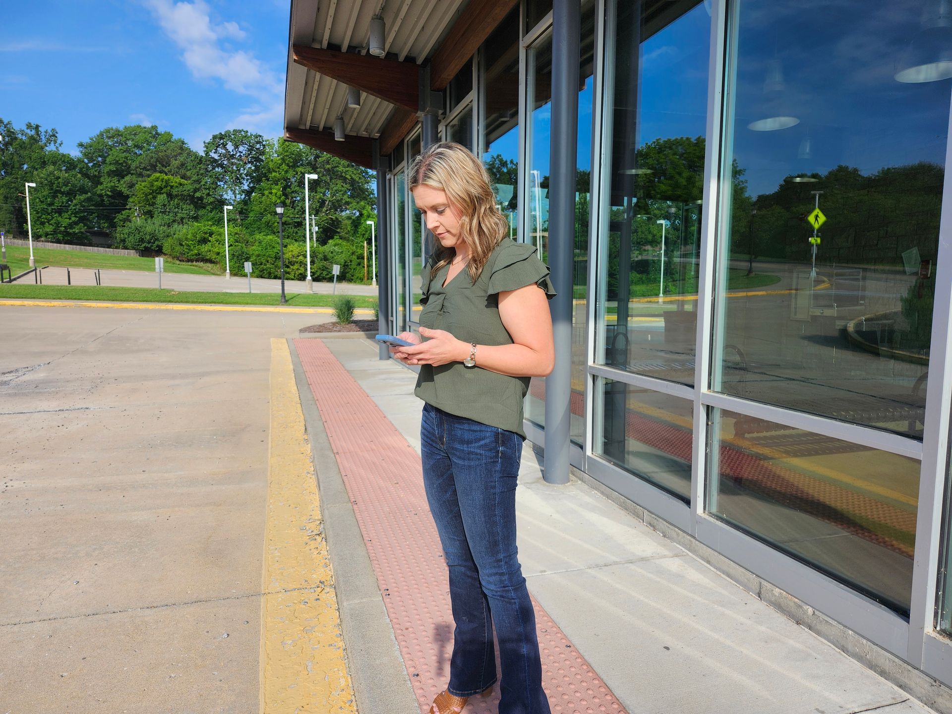 Woman in jeans and green top standing at a bus stop, looking at her phone. Sunny day, tall windows, trees in the background.