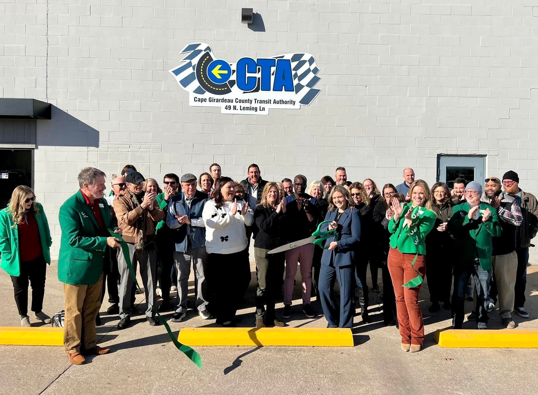 A group of people are standing in front of a building holding a green ribbon.