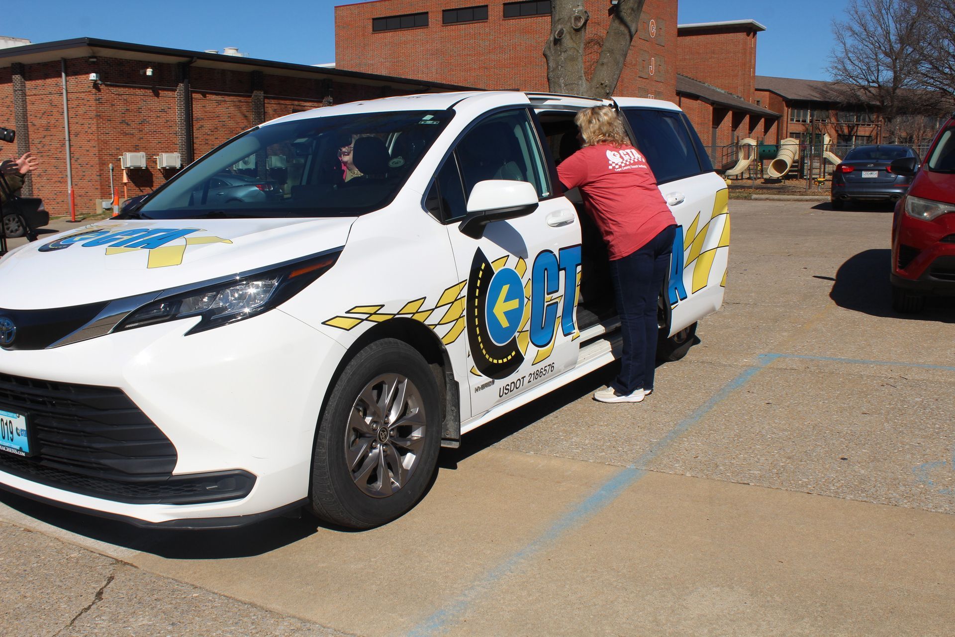 A woman in a red shirt exits a white van with blue logos, parked in a parking lot outside a brick building.