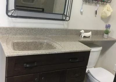 Bathroom vanity with speckled countertop sink, mirror, white toilet, and small wall shelf with decor.