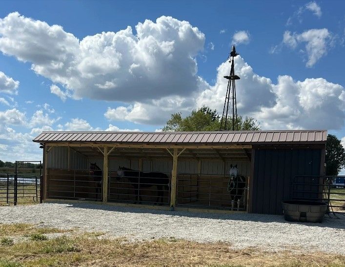 Horses in a shaded barn structure, gravel ground. Brown roof, metal siding. Windmill in the background under a cloudy sky.