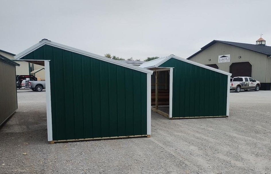 Two dark green sheds with white trim on gravel, background includes trucks and a barn.