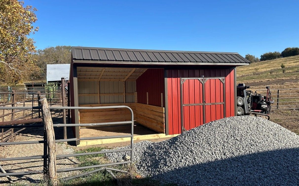 Red shed with a dark roof and gravel pile outdoors, next to a fence under a blue sky.