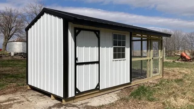 A white chicken coop with a black roof is sitting in the middle of a field.