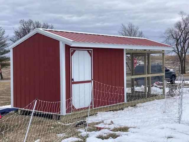 A red shed with a white door is sitting in the snow next to a chicken coop.