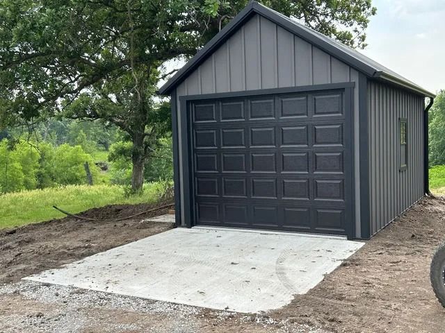 A garage with a black garage door is sitting in the middle of a dirt field.