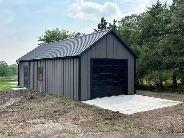 A gray garage with a black garage door is sitting in the middle of a dirt field.