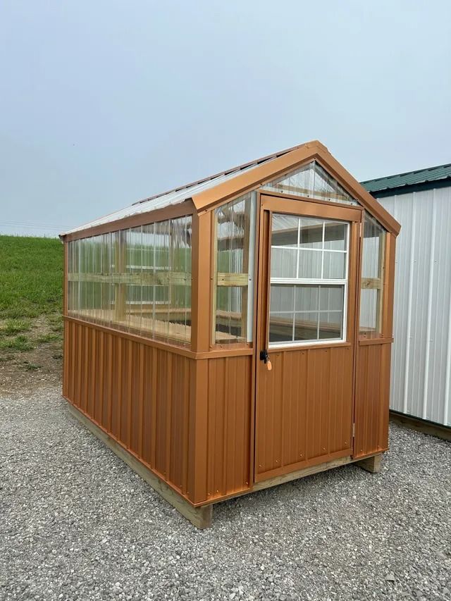 A greenhouse is sitting on top of a gravel lot next to a white building.