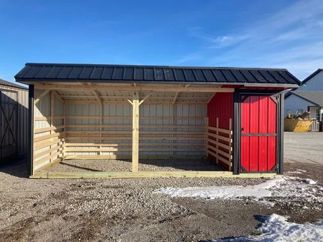 A shed with a red door and a black roof is sitting in the dirt.