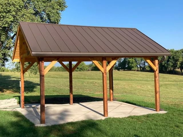 A wooden pavilion with a brown roof is in the middle of a grassy field.