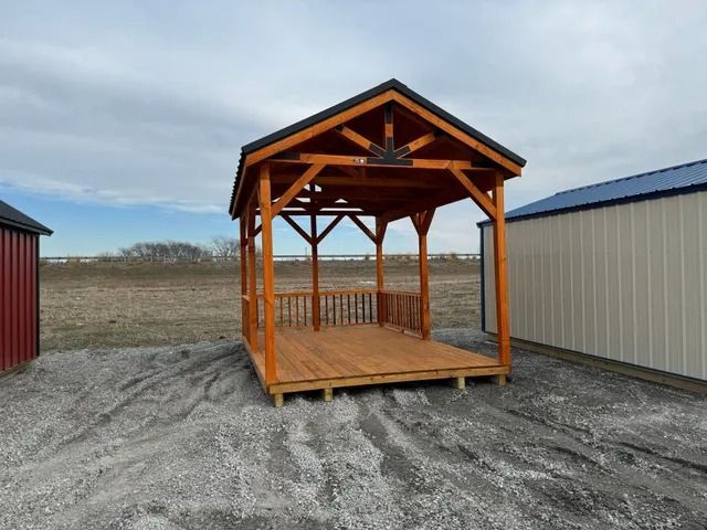 A wooden gazebo is sitting in the dirt in front of a shed.