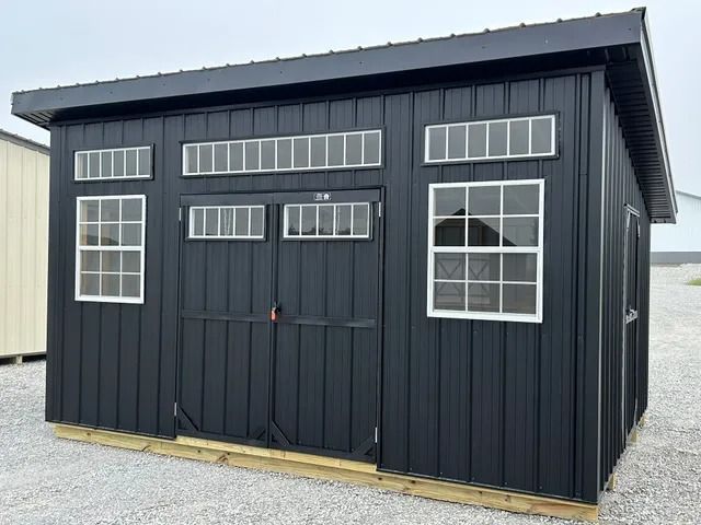A black shed with white windows and doors is sitting on top of a gravel lot.
