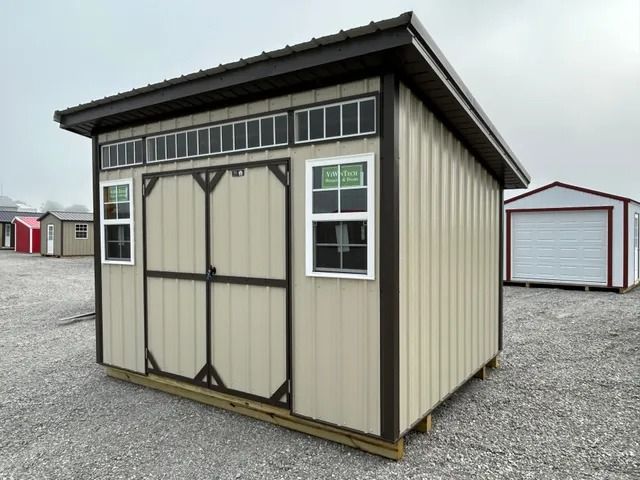 A tan shed with a brown roof and two windows is sitting on top of a gravel lot.