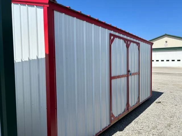 A red and white shed is sitting in a gravel lot next to a building.