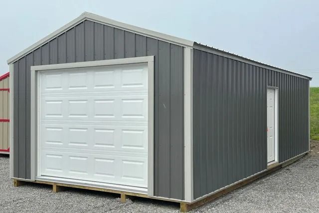 A gray garage with a white garage door is sitting on top of a gravel lot.