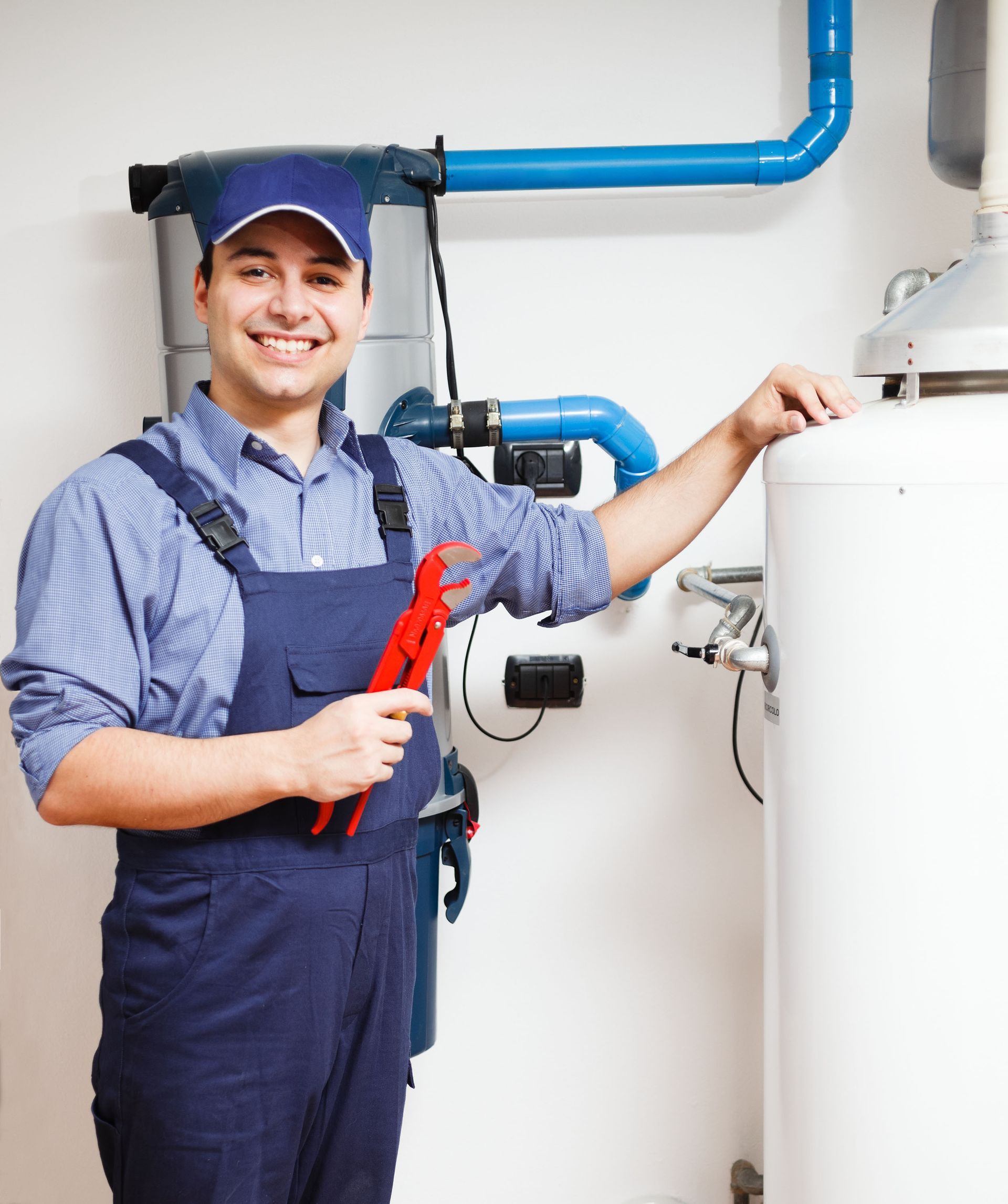 Plumber in blue overalls, holding a red wrench, smiling near a water heater and pipes.