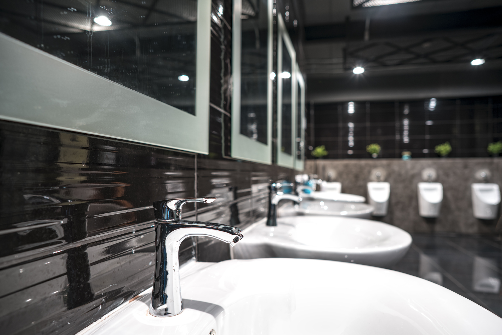 A row of white ceramic sinks with chrome faucets in a modern, dark-tiled public restroom.