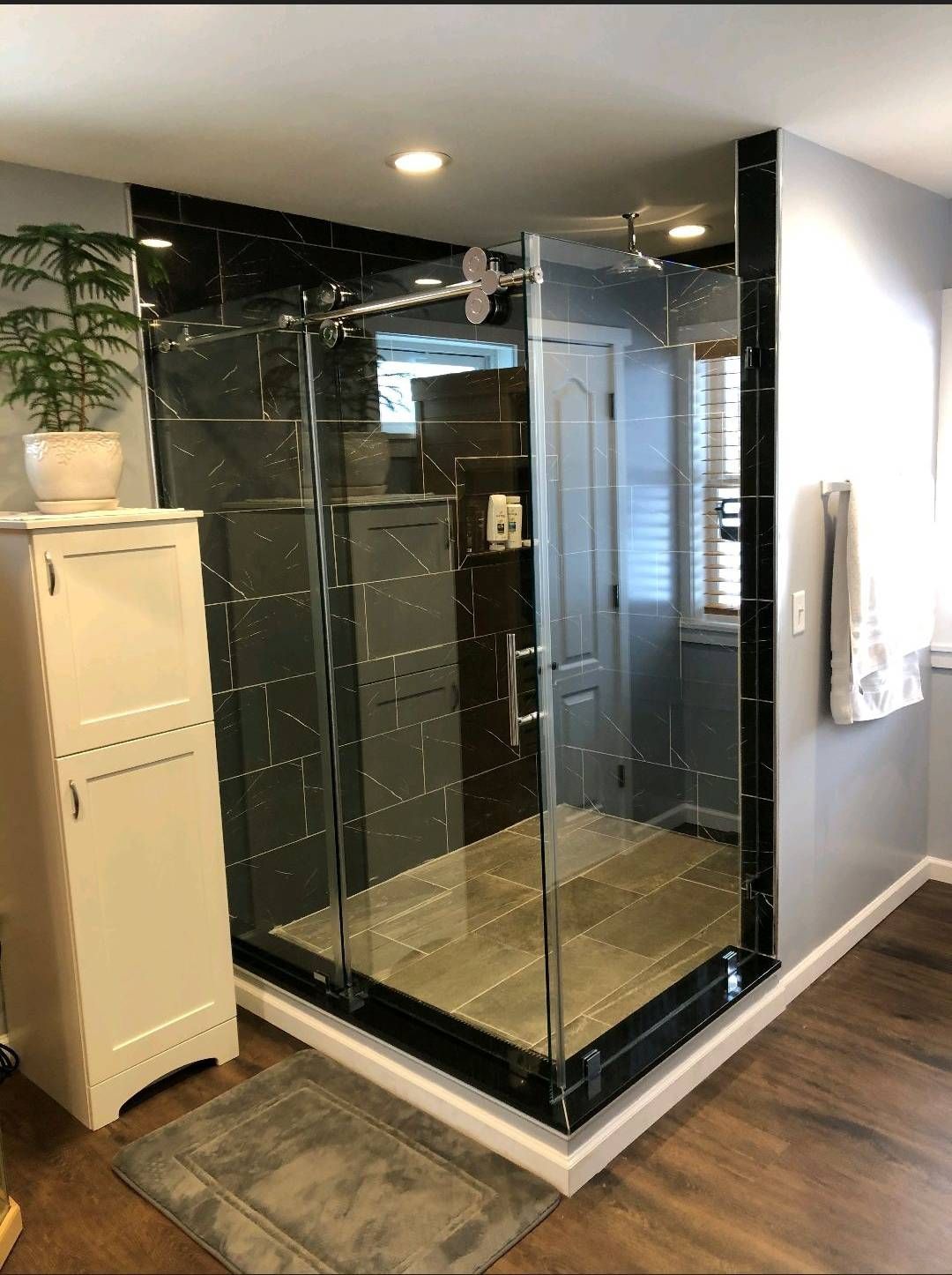 Modern bathroom with glass shower enclosure, black tile, wooden floor, and white cabinet.