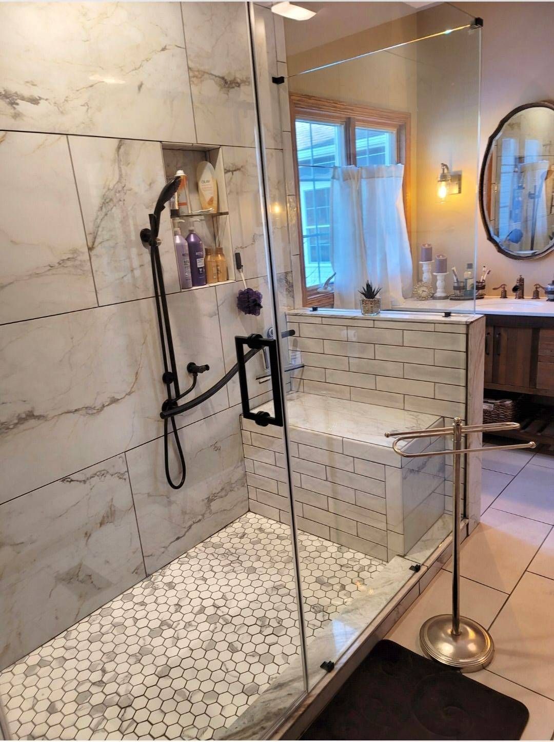 Shower with white tile walls and floor, glass door, built-in bench, black showerhead, and window in background.