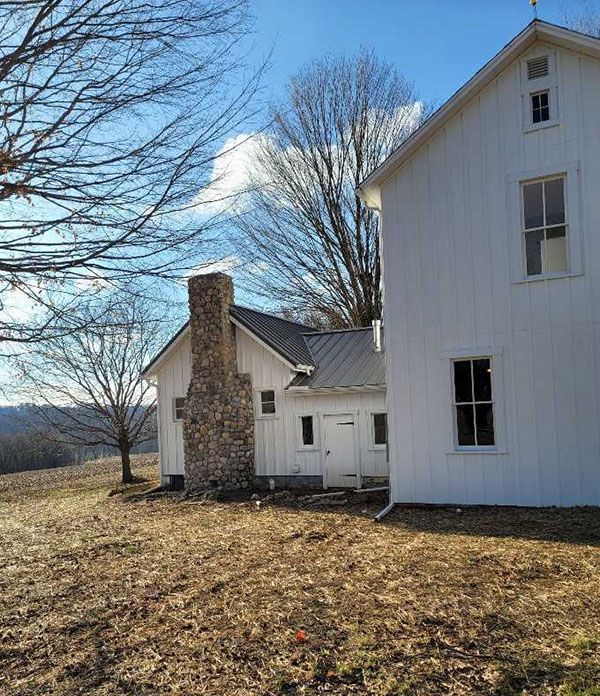 White farmhouse with stone chimney, set in a field under a blue sky, leafless trees.