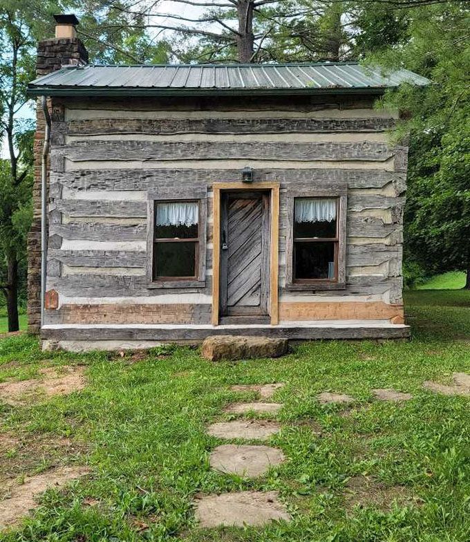 Small weathered log cabin with stone chimney, wooden door, and two windows. Green grass in front, stepping stones leading to entrance.