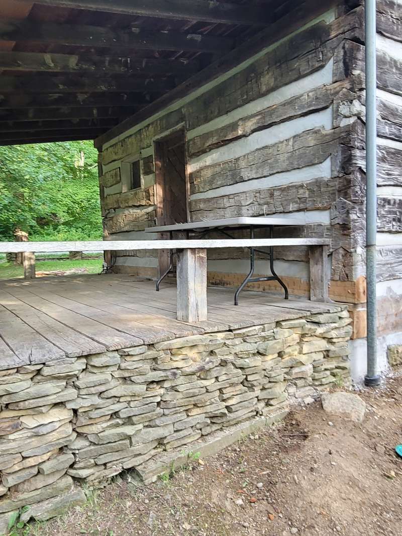Log cabin exterior with stone foundation, porch, and partial view of greenery.