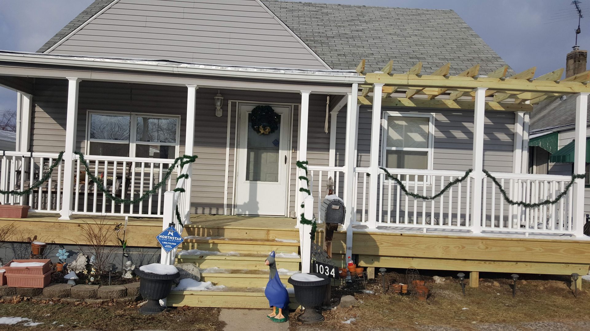 A one-story house with a white porch decorated for the holidays; snow on the ground.