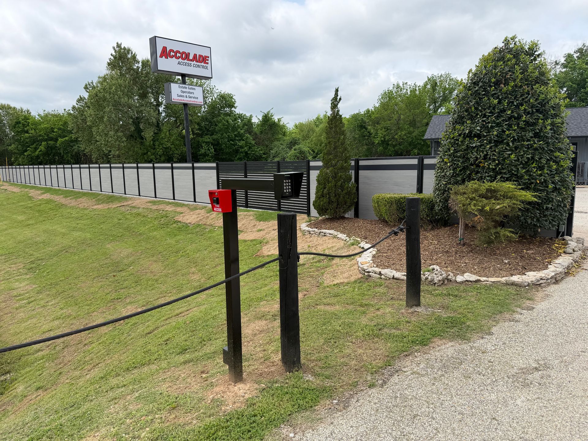 Mailbox and sign beside a gravel driveway, with a fence, grass, and trimmed shrubs under cloudy sky.