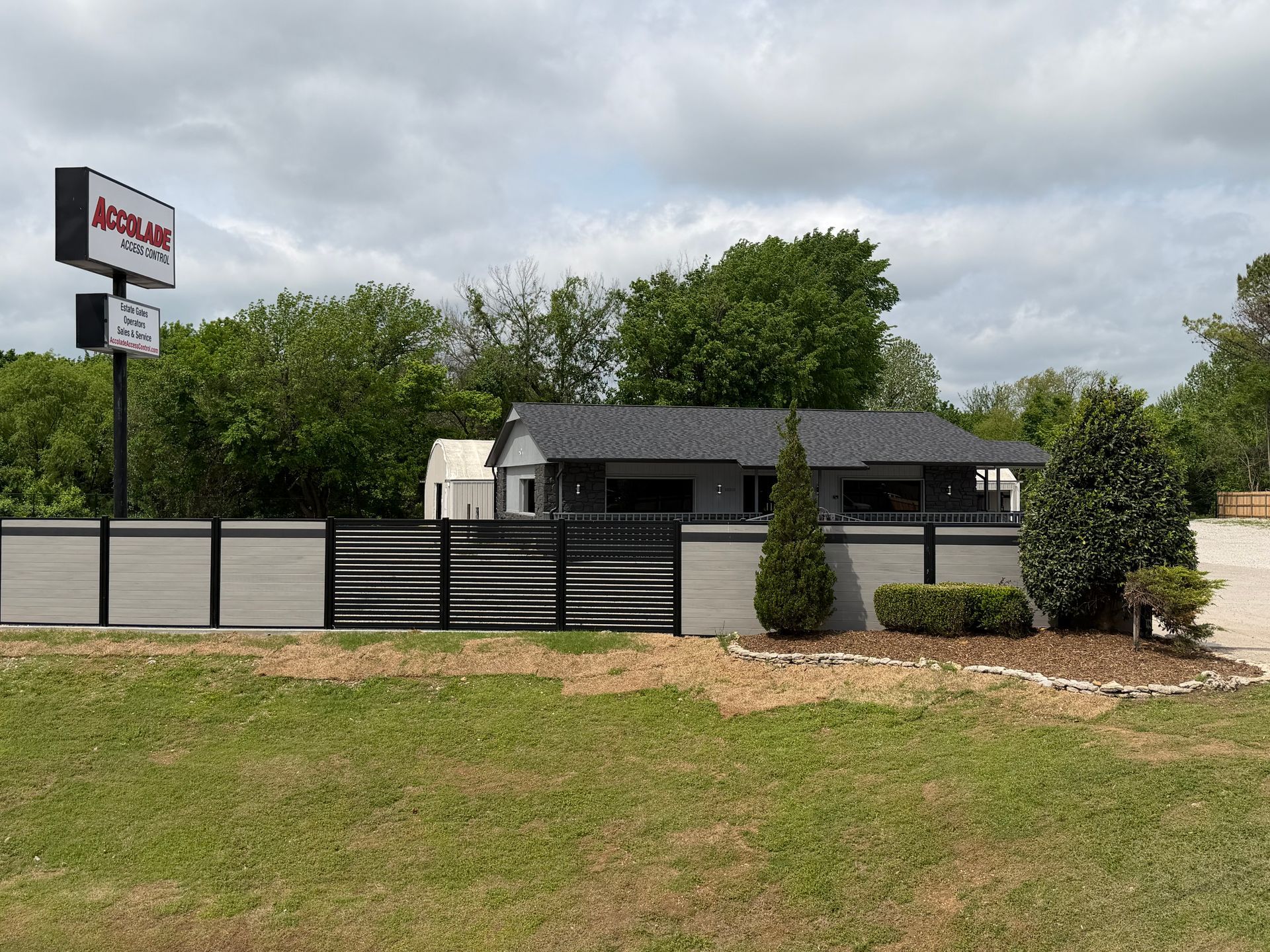Small white building with black roof and fenced yard beside a roadside sign under cloudy skies